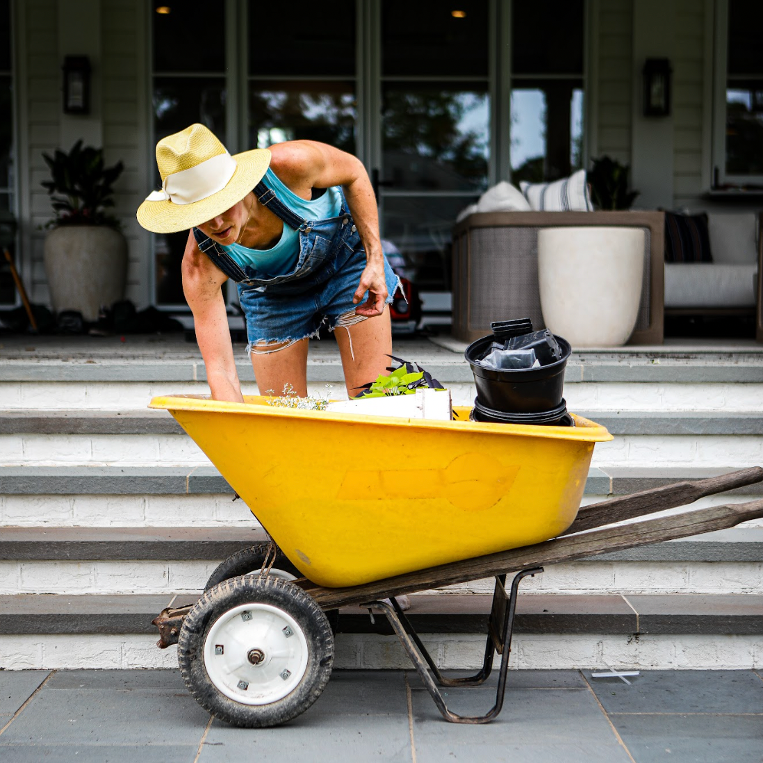 Gardener in yellow hat and overalls reaching in a yellow wheelbarrow in front of patio.