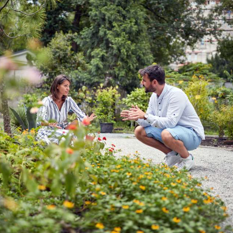 Man and woman bending down talking in garden with green flowers.