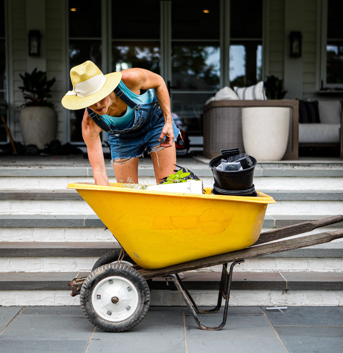 Gardener in yellow hat and overalls reaching in a yellow wheelbarrow in front of patio.